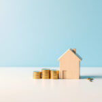 Maison miniature en bois à côté de piles de pièces dorées et d'une clé, sur fond bleu clair