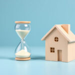 Sablier en verre et maison miniature en bois clair posés côte à côte sur fond bleu clair